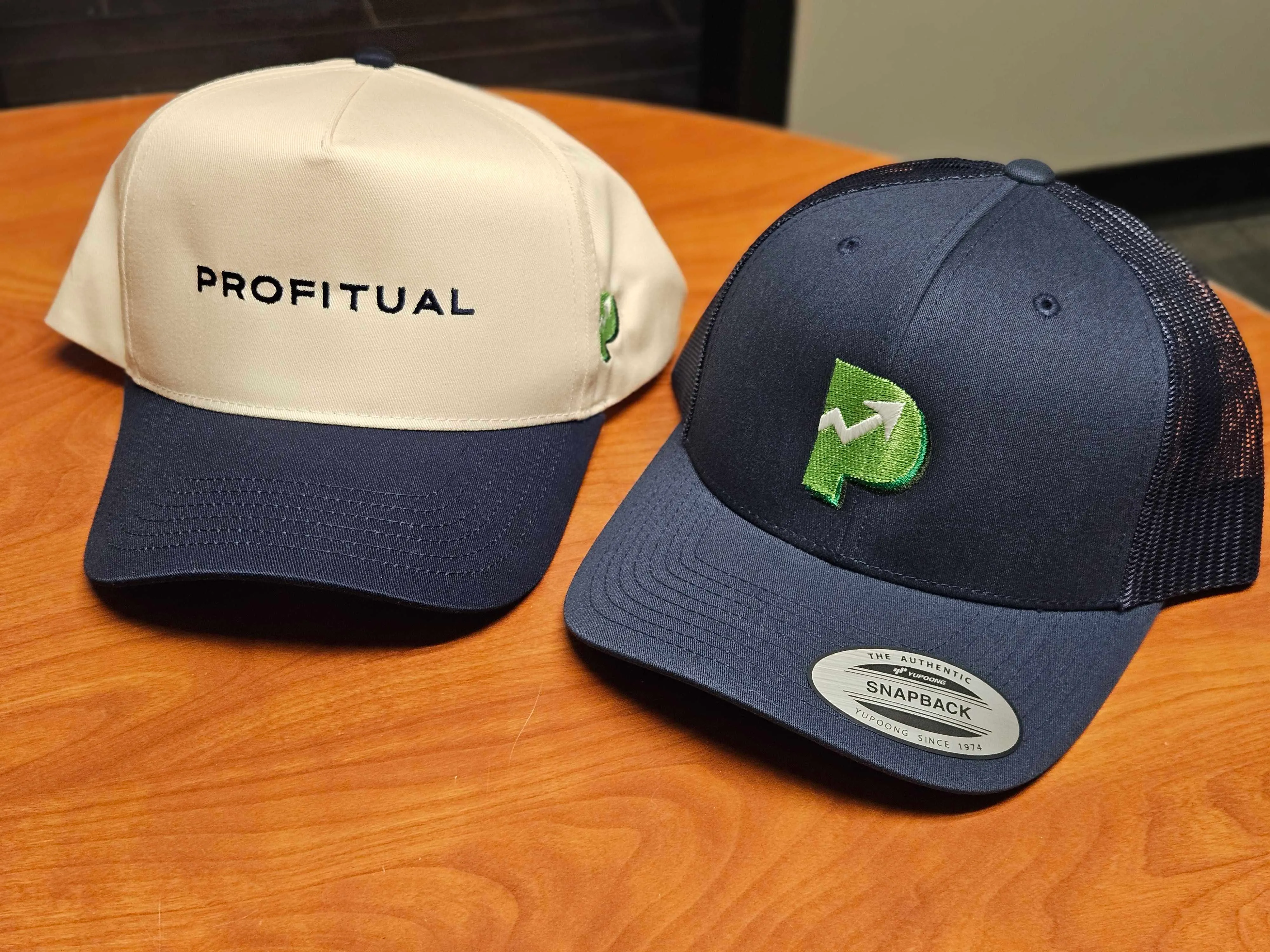 Two Profitual caps — a cream 5-panel with the PROFITUAL wordmark and a navy trucker snapback with the green P logo